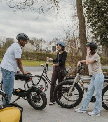 3 jongeren met fietshelm op staan naast hun fietsen en brommer en zijn in gesprek met elkaar.