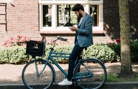 Een man zit op zijn fiets en staat met 1 been op de stoep met een telefoon in zijn hand