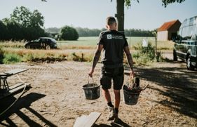 Een man met een shirt van Van Den Bosch metselwerken loopt op een bouwplaats met in iedere hand 1 emmer