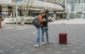 Man en vrouw staan met koffie in de hand en bagage op een stationsplein en kijken op de telefoon die de man vasthoudt