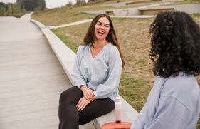 Een vrouw zit op de betonnen rand van een perk en lacht uitbundig naar een andere vrouw, met wie ze in gesprek is.