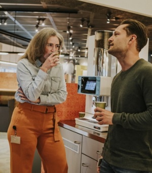 Een man en vrouw staan bij een koffieapparaat en drinken samen een kopje koffie.