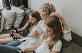 Een vrouw zit op de bank tussen haar beide dochters in. Ze heeft een witte mok vast, en de dochters kijken op hun mobiele telefoons.